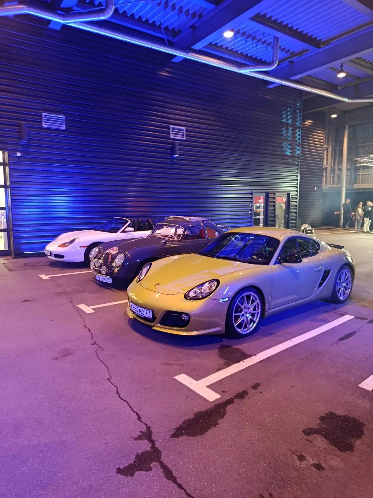 Белый Boxster, тёмный 356 и золотистый Cayman в синем свете у сервисной зоны