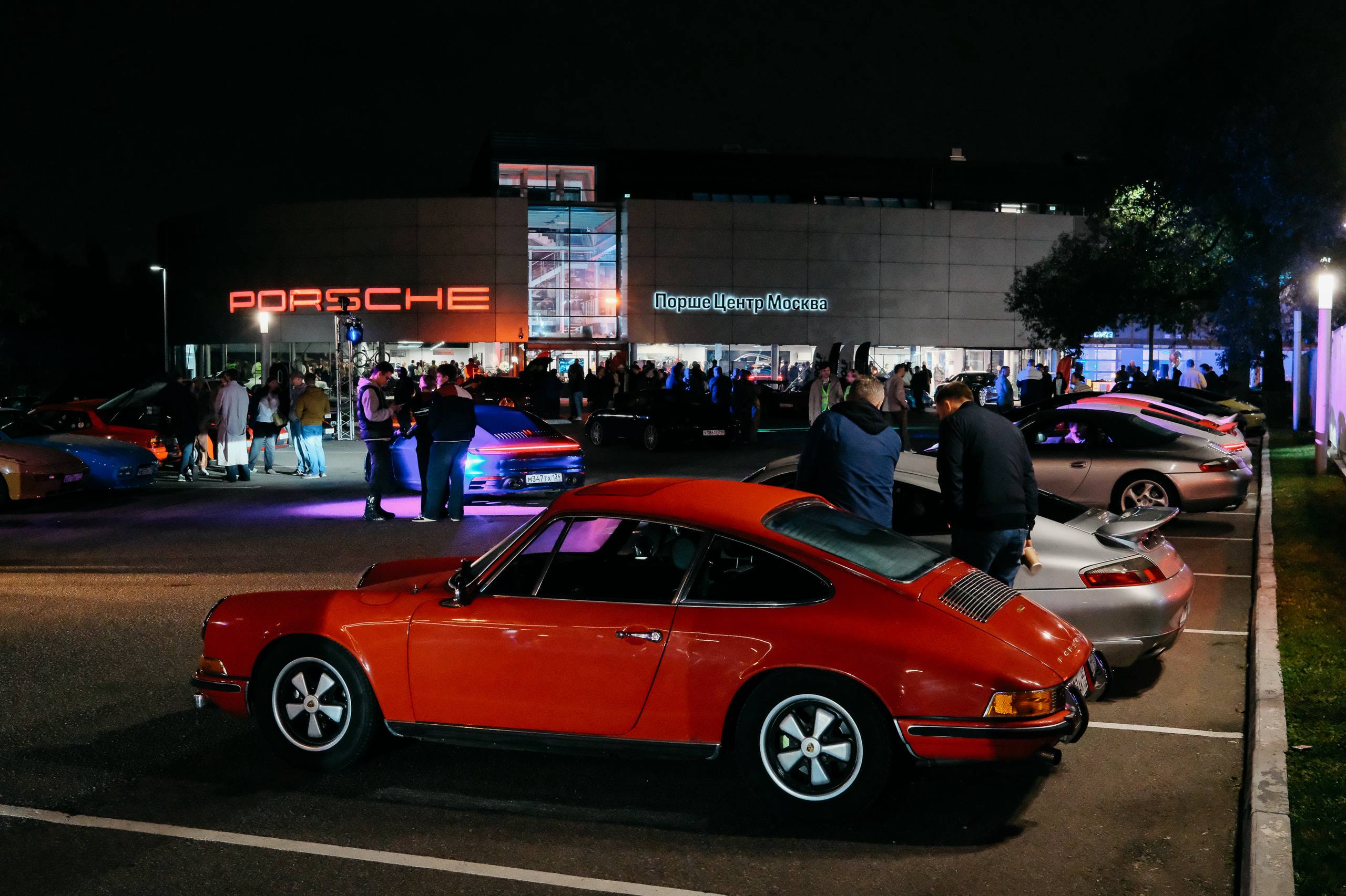 Оранжевый 911 первого поколения на парковке, за ним гости и здание центра с надписью PORSCHE