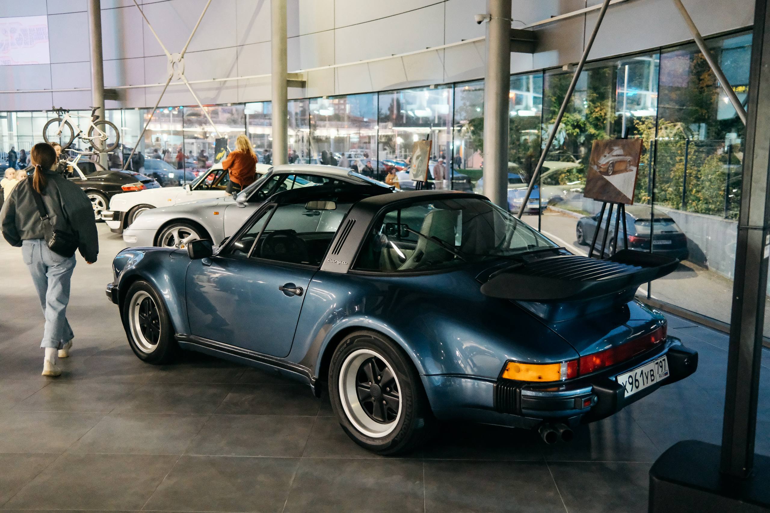 Синий 911 Targa с антикрылом в шоуруме, рядом картины на мольбертах и гости