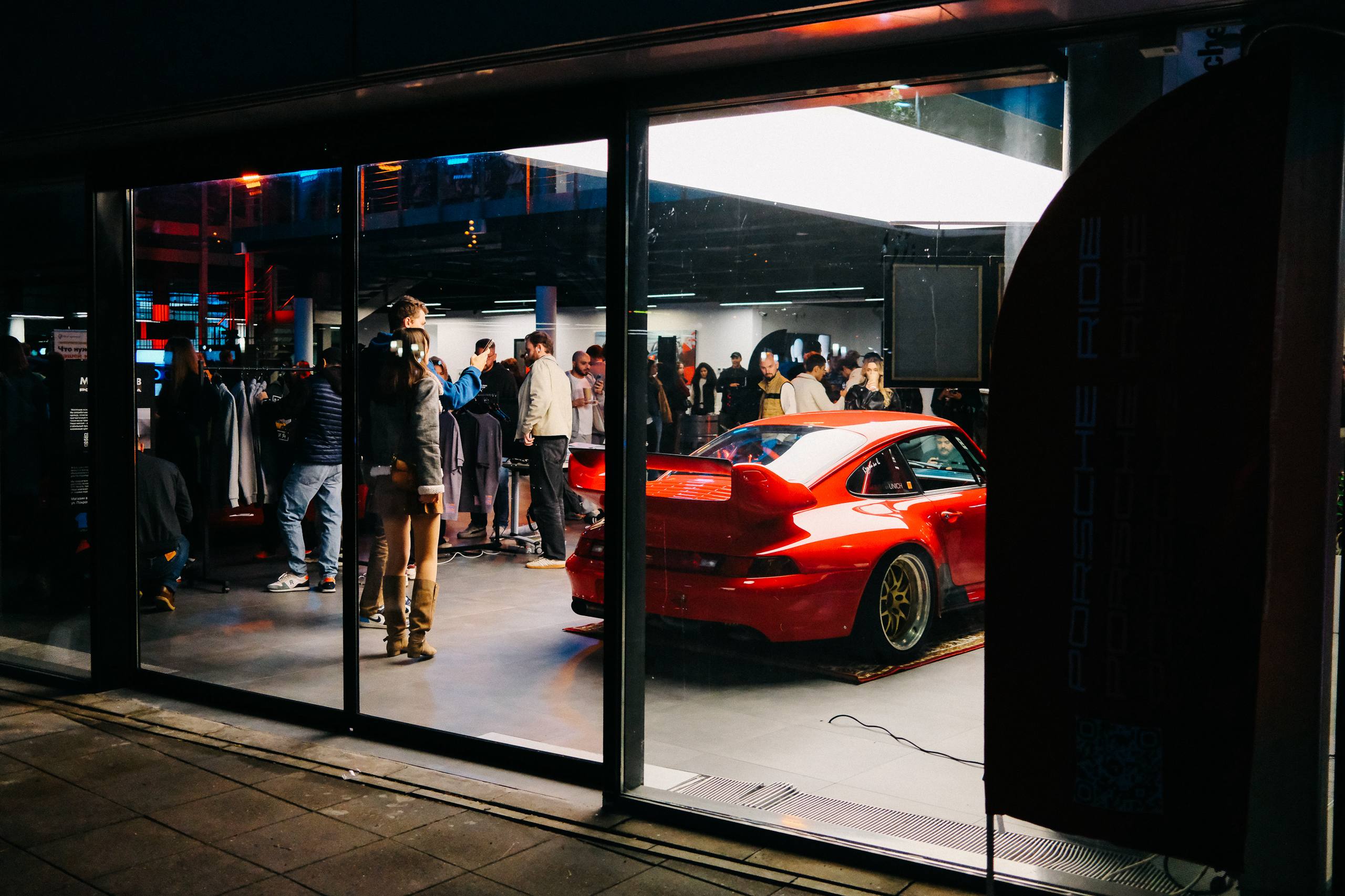 Вид с улицы через витрину: красный 911 (993), гости внутри и флаг Porsche Ride