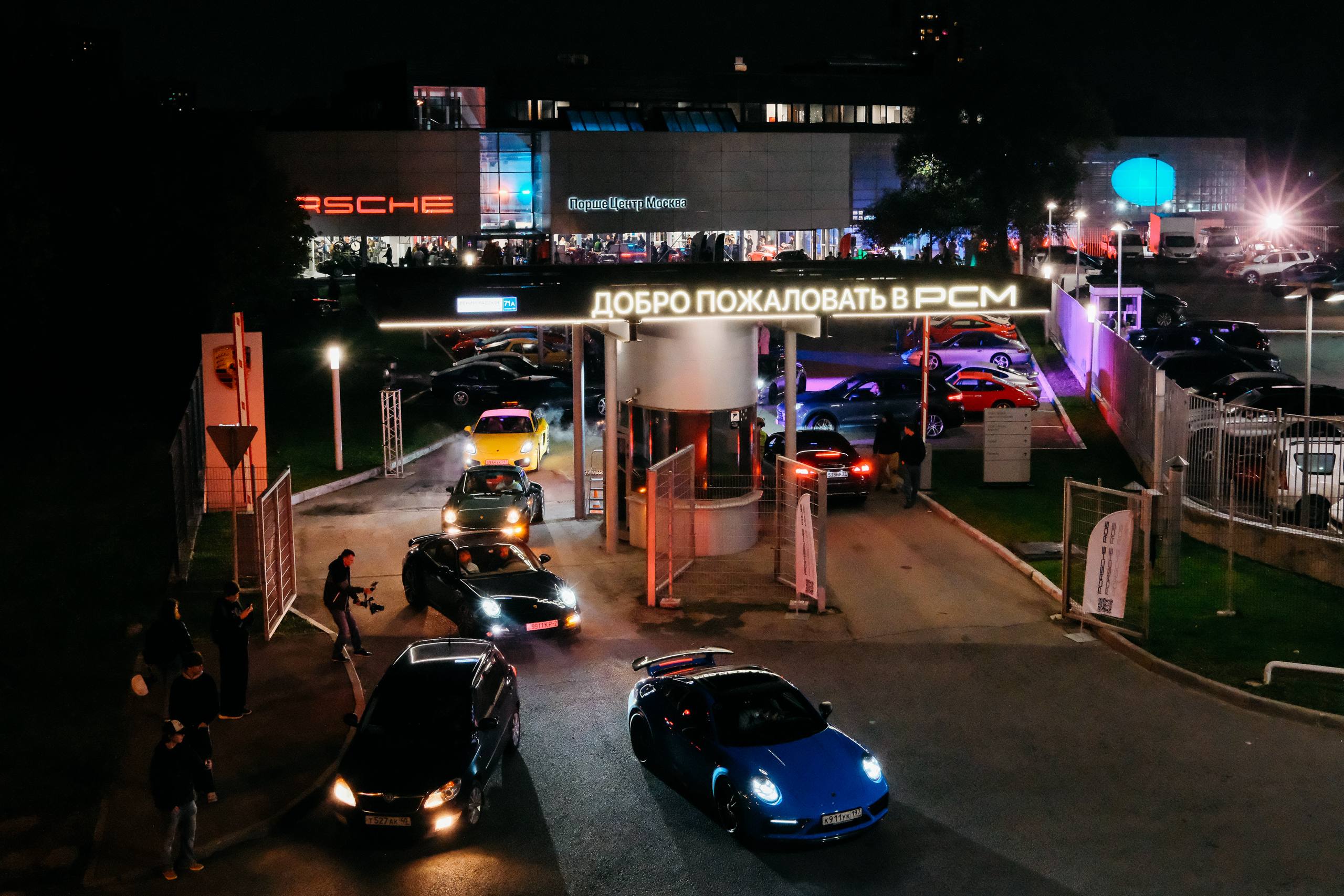 Въезд на территорию PCM ночью: вывеска «Добро пожаловать в PCM», очередь Porsche и гости